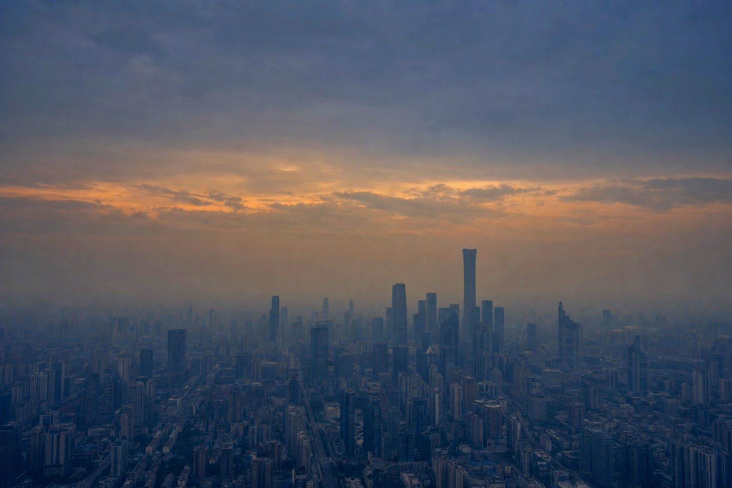 A dense city skyline seen from high above at dusk, a band of amber cloud over hazy blue grey towers
